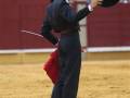 Un torero en el ring, con un sombrero en la mano izquierda y una espada en la otra. El fondo muestra una pared roja con personas observando el evento.