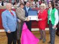 Una celebración en un entorno de toros, con un hombre en traje azul y corbata roja entregando un reconocimiento a una mujer vestida de rojo y con un vestido rosa. Otros individuos observan la escena.