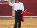 Un torero en el campo de toros, con un traje tradicional y un capote rojo en la mano, preparándose para la corrida de toros.