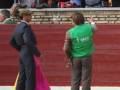 Un torero con un traje azul y un capote rosa se prepara para enfrentarse a una toro en el ruedo de la plaza de toros. Un hombre con un chaleco verde y una camisa blanca sostiene el capote mientras observa la escena. La audiencia, incluyendo personas vestidas con trajes y una mujer en un vestido rojo, observa el evento.