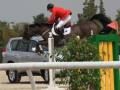 Un jinete monta un caballo negro en una prueba ecuestre, saltando sobre un obstáculo amarillo y verde. El jinete lleva una chaqueta roja, pantalones blancos y un casco negro. La imagen también muestra una parte de un coche blanco detrás del obstáculo.