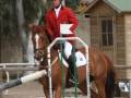 Una mujer monta a un caballo marrón en una pista de equitación, con un arco de obstáculo a su lado. El caballo y la jinete están listos para un salto, con el jinete usando una chaqueta roja y un casco. La imagen muestra la preparación para un evento de salto ecuestre, con el fondo incluyendo una estructura de madera y árboles.