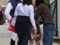 Una persona con una chaqueta de cuadros y botas negras charla con otra en un entorno urbano. La tercera persona, vestida de color marrón y jeans, mira hacia el suelo. En el fondo, se observan un edificio amarillo con techo de ladrillos y una puerta negra.