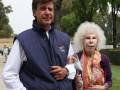 Un hombre y una mujer anciana posan juntos en un parque. El hombre lleva una chaqueta azul con un logotipo y la mujer tiene una bufanda colorida. Ambos están sonrientes, con árboles y césped al fondo.