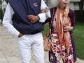 Una pareja caminando por un camino de piedra, el hombre lleva una chaqueta azul con un logotipo y pantalones blancos, mientras la mujer lleva una falda floral y sandalias de tacón.