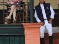 Una anciana con vestido floral y gafas de sol se levanta desde un balcón, mientras un hombre en ropa deportiva y botas de montar observa desde abajo.