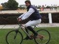 Un hombre en ropa de trabajo blanco y azul monta una bicicleta en un campo verde. En el fondo, se pueden ver carreras de caballos y una estructura de tierra.