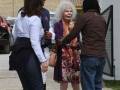 Una mujer con una máscara de terror y vestido floral se encuentra entre dos personas en un entorno urbano con vehículos y estructuras de hormigón.