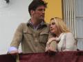 Un hombre y una mujer están mirando hacia el frente desde un balcón con cortinas rojas. El hombre lleva una chaqueta beige y el hombre tiene un cuello de camisa azul. La mujer lleva una blusa blanca con un escote en V y un cuello de camisa. El fondo muestra una pared blanca con un panel amarillo y una ventana con persianas.