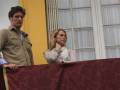 Un hombre y una mujer se encuentran en un balcón con cortinas blancas. El hombre está de pie, mirando hacia el frente, mientras la mujer se inclina ligeramente hacia adelante. Ambos están vestidos formalmente, lo que sugiere una ocasión especial o formal. La imagen está tomada en un entorno urbano, con una pared amarilla visible detrás de ellos.