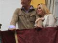 Un hombre y una mujer están en un balcón, el hombre sostiene una bandera roja con detalles dorados. Ambos sonríen y parecen estar en un evento formal o festivo.