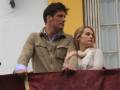 Un hombre y una mujer miran hacia el horizonte desde un balcón con cortina roja.