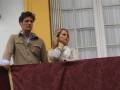 Dos personas, uno hombre y una mujer, están en un balcón con cortinas blancas. El hombre lleva una chaqueta beige y la mujer tiene el cabello largo y se viste con un abrigo blanco. La imagen parece ser de una celebración o evento formal, dado el estilo de vestimenta y la decoración del balcón.