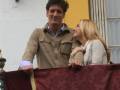 Un hombre y una mujer sonríen mientras se apoyan en un balcón con una cortina roja, frente a una fachada de edificio blanco y amarillo.