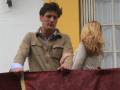 Un hombre y una mujer están en un balcón con una cortina roja. El hombre tiene el cabello oscuro y lleva una chaqueta beige, mientras que la mujer tiene el cabello largo y rubio. La imagen muestra un ambiente urbano con una pared blanca al fondo y una ventana con persianas.