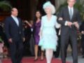 Una anciana elegante con vestido azul menta y sombrero azul se desplaza por un pasillo decorado con plantas tropicales. A su lado, dos hombres en trajes de negocios y una mujer en vestido morado caminan por un pasillo con alfombra roja.