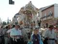 Procesión religiosa con estandarte ornado y participantes en trajes tradicionales, en una calle urbana.