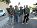 Cuatro hombres con camisetas verdes y sombreros de ala ancha, posando en una calle con un coche blanco y un tractor en el fondo.