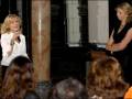 Una mujer habla en un micrófono mientras otra observa, frente a una columna de mármol. La escena parece ser en un evento formal o conferencia.