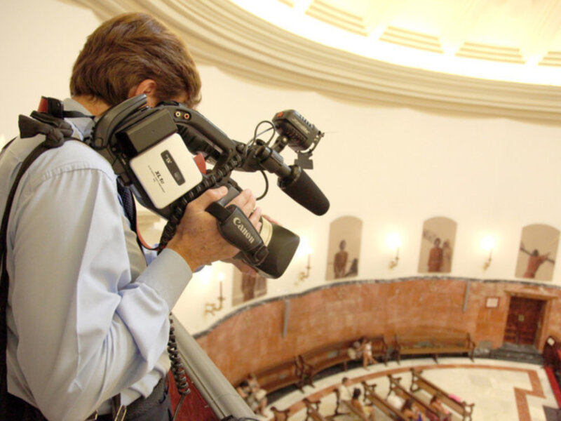 Un hombre con una cámara de video en un interior iluminado, probablemente un edificio público o institucional. La cámara está montada en una trípode y el hombre parece estar grabando o filmando algo. La imagen muestra un ambiente con una arquitectura clásica, con columnas y estatuas en el fondo.