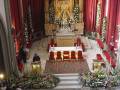 Interior de una iglesia con altar dorado, flores y velas. Una persona en el púlpito hablando a una audiencia sentada en sillas rojas. Tapices y cortinas rojas adornan el espacio.