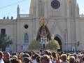 Fotografía de una procesión religiosa frente a un edificio histórico con una imagen venerada en el centro. La imagen muestra a una multitud de personas observando la procesión, mientras que algunos están en las alturas del edificio. El cielo es claro y el ambiente parece ser una celebración o evento importante en la comunidad local.