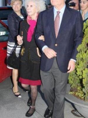 Una pareja camina juntos por una calle, el hombre lleva un traje azul y la mujer una chaqueta negra con detalles rojos. Detrás de ellos, se pueden ver edificios y árboles.