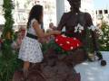 Una figura de chocolate representa a una mujer con un vestido negro y un tocado. La escultura está decorada con flores de chocolate y tiene una mano extendida hacia un vestido blanco. La escena se desarrolla en un entorno con columnas y plantas, posiblemente en una celebración o evento.