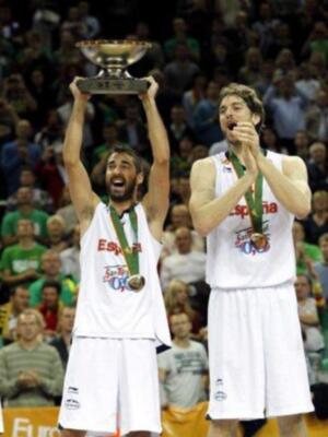Dos jugadores de baloncesto celebran con trofeos y medallas, uno levantando un trofeo grande. El fondo muestra a una multitud de espectadores en un estadio lleno, con una camiseta visible que dice "España" y el año 2013.