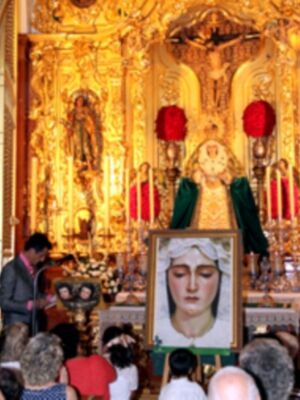 Interior de una iglesia con altares dorados y estatuas religiosas. En el centro, una imagen de la Virgen María con un niño en sus brazos. A su lado, una estatua de la Virgen María con un niño en sus manos. En el fondo, un altar dorado con velas y adornos. Una persona habla en una tribuna de madera, frente a un grupo de personas que observan la imagen.