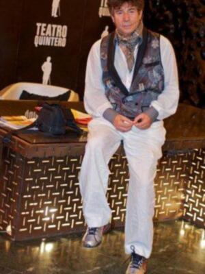 Un hombre se sienta en un banco de madera con patrones, vestido con una chaqueta estampada, pantalones blancos y zapatillas deportivas. En el fondo, hay un cartel con la palabra "Teatro Quinteros" y una figura de un actor.