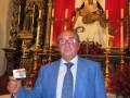 Un hombre en traje azul posa frente a una imagen religiosa en un altar dorado, rodeado de flores rojas y velas. La imagen muestra una escena de devoción y respeto a la religión.