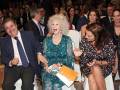Una celebridad en un evento formal, sentada entre otros invitados. La imagen muestra a una mujer con vestido verde y corona, junto a un hombre en traje y corbata. La escena parece ser una gala o conferencia, con asistentes en el fondo.