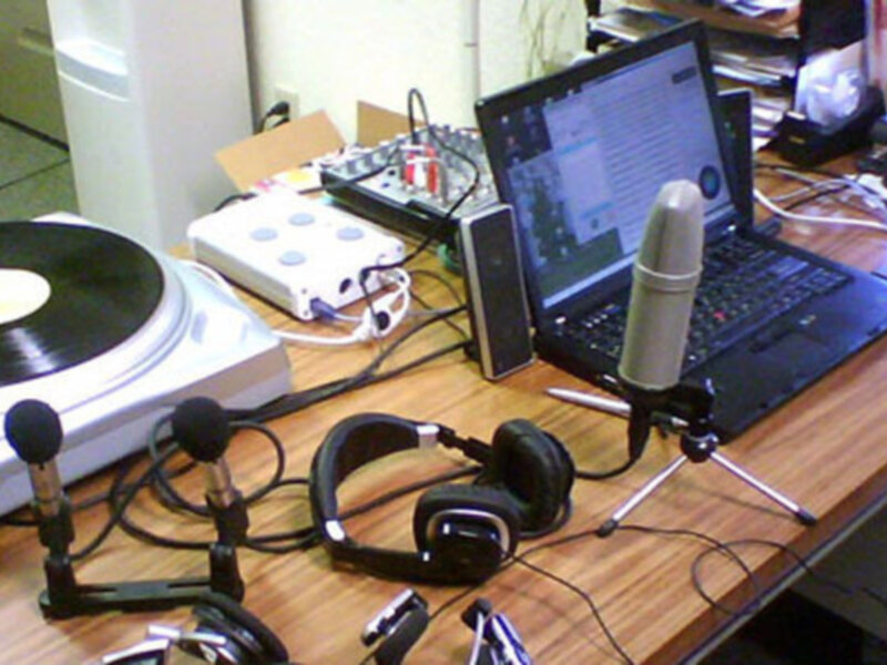 Un escritorio con un portátil, auriculares, micrófonos y un vinilo giradiscos, preparado para grabar o transmitir audio en vivo.
