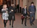 Una imagen de cuatro personas caminando por una calle iluminada, dos mujeres con chaquetas negras y tacones altos, una mujer con botas de cuero y una persona masculina en abrigo azul.