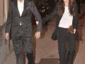 Un hombre y una mujer caminan juntos por la calle, ambos vestidos con trajes formales. La mujer lleva un vestido blanco y pantalones negros, mientras que el hombre usa un traje negro con una camisa blanca. Ambos sonríen y tienen sus manos entrelazadas, lo que sugiere una relación romántica. La imagen captura un momento de felicidad y estabilidad en el ambiente urbano.