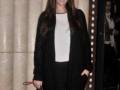 A una mujer con cabello largo y oscuro, vestida de negro con un blazer y pantalones. Lleva una cartera con estampado de leopardo en la mano izquierda y sonríe. La escena parece ser una alfombra roja o evento formal, con cortinas y luces de fondo.