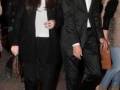 Dos personas caminan juntos en un ambiente nocturno, uno con traje negro y la otra con abrigo largo y blusa. La imagen sugiere una ocasión formal o social.