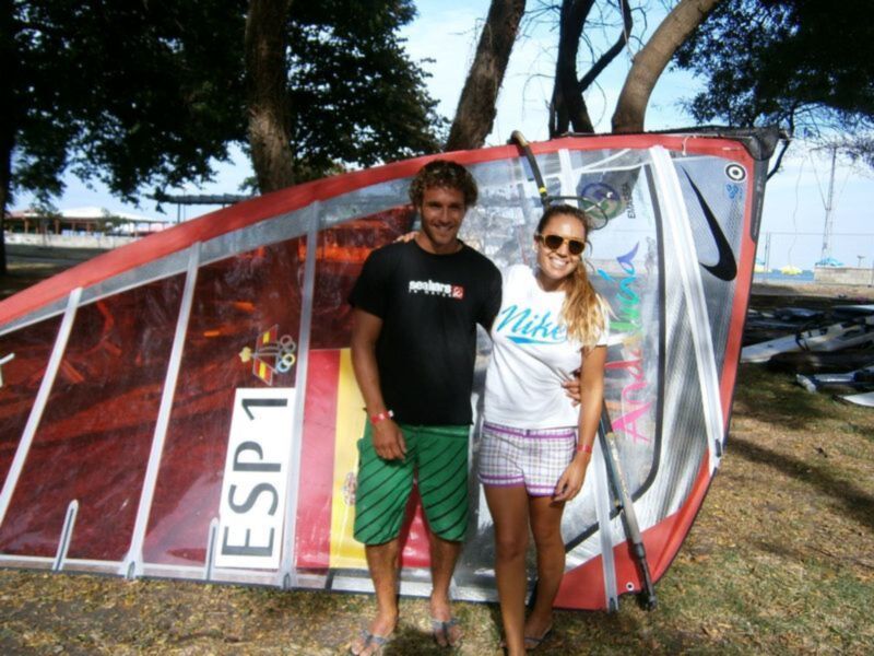 Dos personas posan junto a una vela de windsurf en un entorno al aire libre.