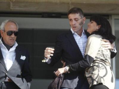Una persona en un traje formal sostiene la mano de otra persona con una chaqueta estampada. Ambos están en un entorno urbano, posiblemente una calle o pasillo de edificio.