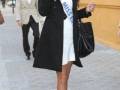 Miss España 2012 caminando por la calle, con elegante traje negro y vestido blanco, sosteniendo una maleta negra.