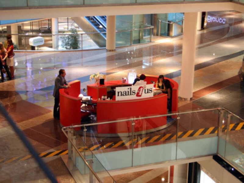 Puesto de manicura 'nails4us' en un centro comercial con escaleras y tiendas al fondo.