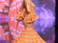 Una modelo presenta un vestido de flamenco naranja con volantes en la falda y mangas con volantes en un desfile de moda.