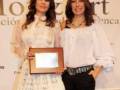 Dos mujeres sonrientes sostienen un reconocimiento en un evento formal. La imagen está acompañada por una bandera que parece ser de la marca "Mozart".