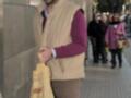 Un hombre camina por la calle, llevando una bolsa de M&M's en su mano izquierda. Está vestido con un chaleco beige, una camisa de manga larga roja y pantalones marrones. La imagen muestra a otras personas en el fondo, caminando y hablando.