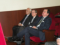 Tres hombres en trajes sentados, uno con los pies sobre el asiento del otro. La imagen parece ser de una reunión o conferencia formal en un entorno interior con paredes de madera y asientos rojos.