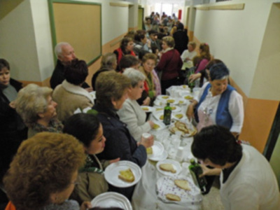 Una larga fila de personas sentadas en mesas, compartiendo comida y conversando. La escena parece ser un comedor o evento social en una sala con paredes blancas y colores.