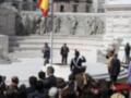 Estatua de un hombre en el centro de una plaza con una bandera en el fondo, mientras una persona toma una foto. El entorno parece ser un monumento o un lugar histórico.