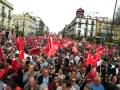 Una multitud en un evento político o sindical, ondeando banderas rojas y blancas. En el fondo, edificios históricos con carteles de propaganda política.