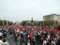 Una multitud de personas con pancartas rojas y banderas ondeando en una manifestación. En el fondo, se pueden ver edificios históricos y un monumento central.